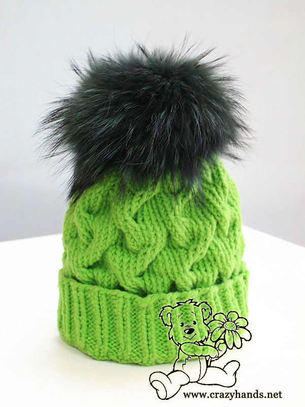finished winter cable hat knit using green yarn with attached dark green fur pom