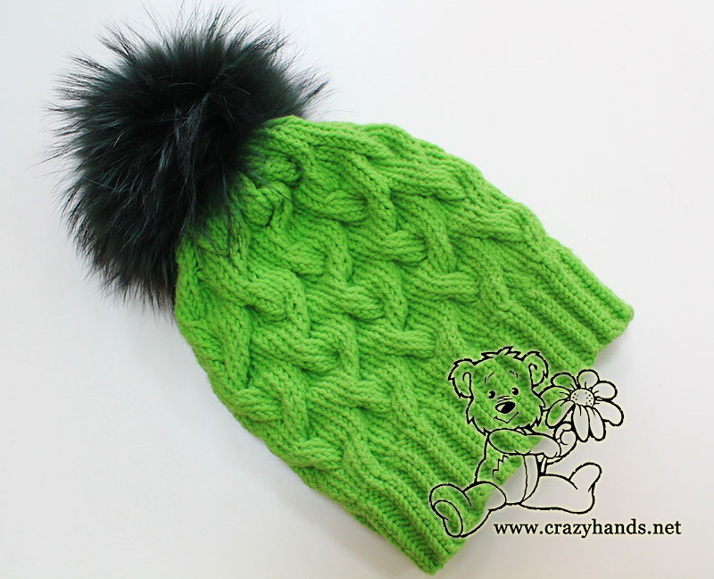 winter cable hat made with green yarn and decorated with dark green raccoon fur pom pom