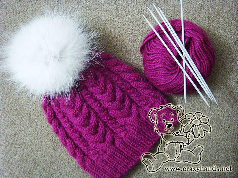 women's cable knit winter hat with white fur pom pom and yarn skein and needles on the side