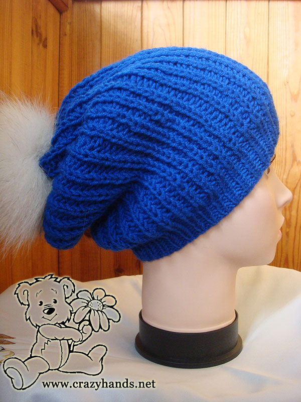 slouchy knit hat made with blue yarn and decorated with white fur pom pom. shot is made on the mannequin's head