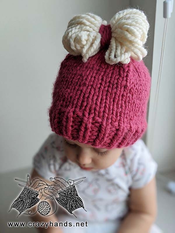baby girl wears a beanie made with super bulky violet yarn and decorated with white yarn bow