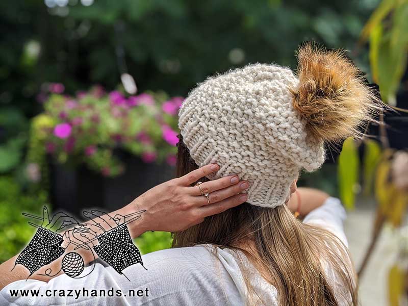 model wears a bulky snowflake knit hat with faux fur pom pom. shot from the back