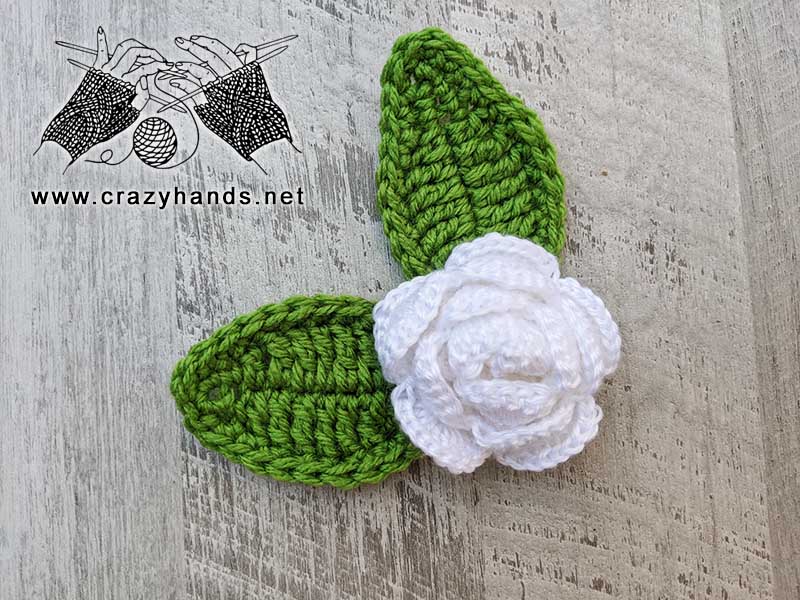 two crochet elliptical leaves next to a white crochet rose forming a flower