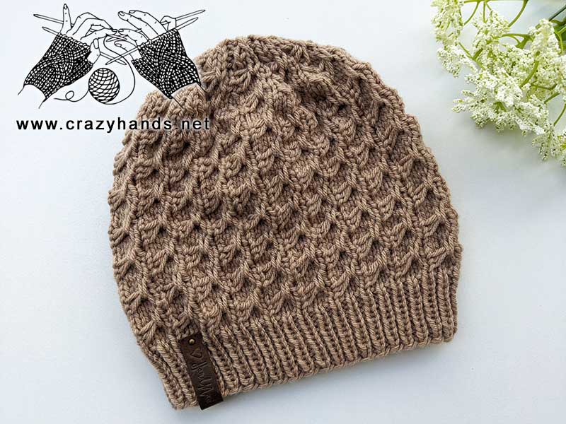 swivel knit winter hat, made with light brown yarn, next to a white flower